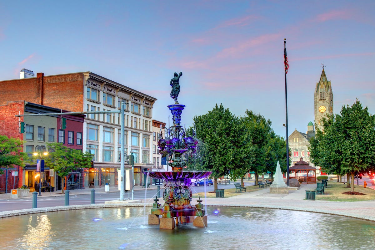 Downtown Watertown, featuring a fountain with a purple statue in the foreground and a small building and a church in the background